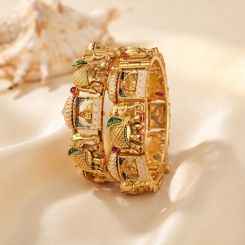A stack of ornate gold-toned bangles with detailed elephant motifs, white stone inlays, and small red and green enamel accents, displayed on a soft cream satin surface with a seashell in the background.