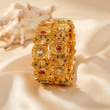 An ornate gold-toned bangle bracelet decorated with red, green, and white stones, displayed on draped cream satin fabric with a seashell in the background.