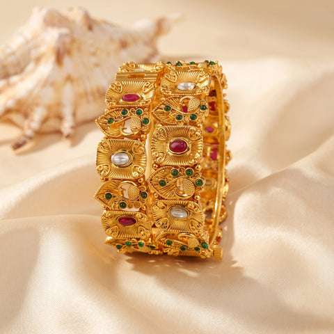 An ornate gold-toned bangle bracelet decorated with red, green, and white stones, displayed on draped cream satin fabric with a seashell in the background.