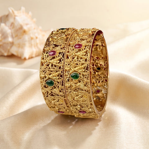 An ornate gold-toned bangle bracelet with intricate filigree patterns and small green and pink gemstone accents, displayed on draped cream satin fabric with a seashell in the background.