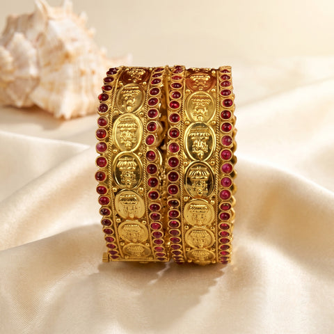 A set of three gold-plated traditional Indian bangles decorated with red gemstone accents, displayed on a cream satin fabric with a seashell prop in the background.