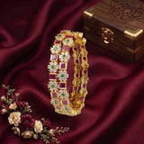 A gold-plated Indian-style bangle bracelet decorated with alternating red, green, and white stones, displayed on a draped dark red satin fabric next to a wooden box and flowers.