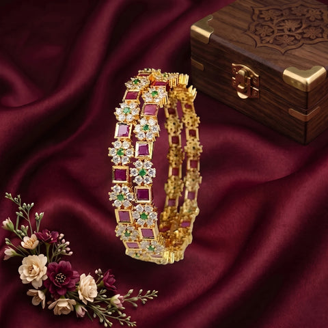 A gold-plated Indian-style bangle bracelet decorated with alternating red, green, and white stones, displayed on a draped dark red satin fabric next to a wooden box and flowers.