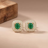 A pair of gold-plated square stud earrings featuring a central green emerald-cut gemstone surrounded by a halo of small clear pavé stones, displayed on a beige surface with a wooden block in the background.