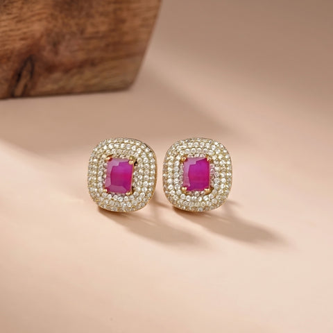 A pair of gold-toned stud earrings with rectangular pink gemstones surrounded by small clear stones, displayed on a warm beige surface with a blurred wooden object in the background.