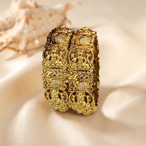 An ornate gold-toned bangle bracelet with intricate filigree patterns and small clear stone accents, displayed on draped cream satin fabric with a seashell in the background.