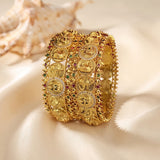 An ornate gold-toned multi-strand bangle bracelet with intricate filigree, floral motifs, and small red and green stone accents, displayed on a soft cream satin surface with a seashell in the background.