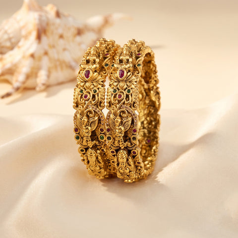 A pair of ornate gold bangles with detailed filigree and small red, green, and black stone accents, displayed on a beige satin surface with a seashell in the background.