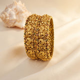 An ornate gold-toned cuff bracelet with intricate floral and scroll motifs, accented by small red and green gemstones, displayed on draped cream fabric with a seashell in the background.