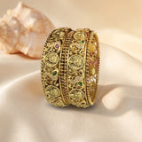 Three ornate gold-toned bangles with intricate filigree and small green and pink stone accents, displayed on draped cream fabric with a seashell in the background.