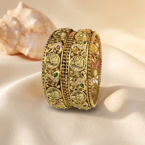 Three ornate gold-toned bangles with intricate filigree and small green and pink stone accents, displayed on draped cream fabric with a seashell in the background.