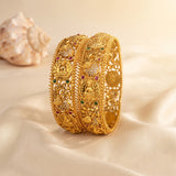 An ornate gold-toned traditional Indian bangle bracelet with intricate filigree, floral motifs, and small red and green stone accents, displayed on a cream satin fabric with a seashell prop in the background.
