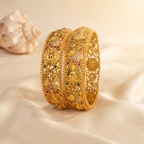 An ornate gold-toned traditional Indian bangle bracelet with intricate filigree, floral motifs, and small red and green stone accents, displayed on a cream satin fabric with a seashell prop in the background.