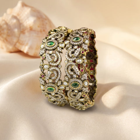 An ornate gold-toned bangle bracelet decorated with green stones, clear rhinestones, and white faux pearls, displayed on a beige satin fabric with a seashell in the background.