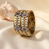 An ornate gold-toned bangle bracelet with intricate silver-toned pavé detailing and oval blue gemstones, displayed on draped cream satin fabric with a seashell in the background.