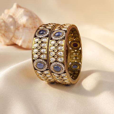 An ornate gold-toned bangle bracelet with intricate silver-toned pavé detailing and oval blue gemstones, displayed on draped cream satin fabric with a seashell in the background.