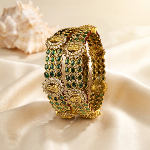 A close-up of an ornate gold-toned bangle bracelet decorated with green stone accents and small clear stones, displayed on draped cream satin fabric with a seashell in the background.