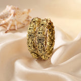 A chunky gold bangle bracelet with intricate 3D owl and bird motifs, displayed on draped cream satin fabric with a seashell in the background.