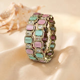 A gold-plated triple-strand bracelet featuring rectangular pastel gemstones in pink, mint green, and purple, accented with clear rhinestones, displayed on a beige satin fabric with a seashell in the background.