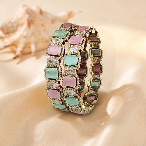 A gold-plated triple-strand bracelet featuring rectangular pastel gemstones in pink, mint green, and purple, accented with clear rhinestones, displayed on a beige satin fabric with a seashell in the background.