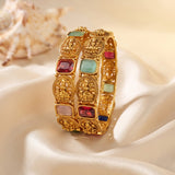 A close-up of a gold-toned bangle bracelet with multiple rectangular and oval inlaid stones in red, light blue, pink, and dark blue, displayed on draped cream satin fabric with a shell in the background.