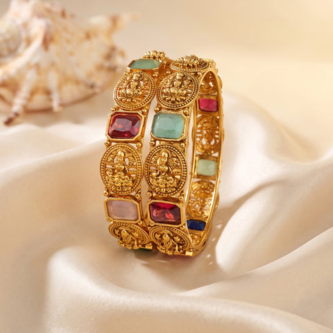 A close-up of a gold-toned bangle bracelet with multiple rectangular and oval inlaid stones in red, light blue, pink, and dark blue, displayed on draped cream satin fabric with a shell in the background.