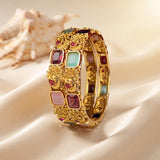 A wide gold-plated bangle bracelet decorated with colorful rectangular and round stones, displayed on draped cream satin fabric with a seashell in the background.