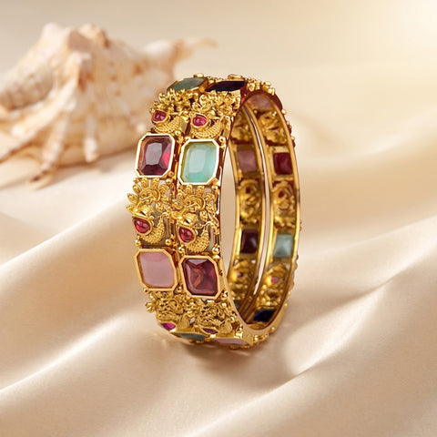 A wide gold-plated bangle bracelet decorated with colorful rectangular and round stones, displayed on draped cream satin fabric with a seashell in the background.