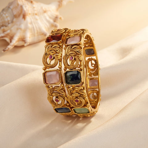 A wide gold bangle bracelet with ornate filigree detailing and multiple rectangular inset stones in pink, dark blue, green, and red, displayed on draped cream fabric with a seashell in the background.