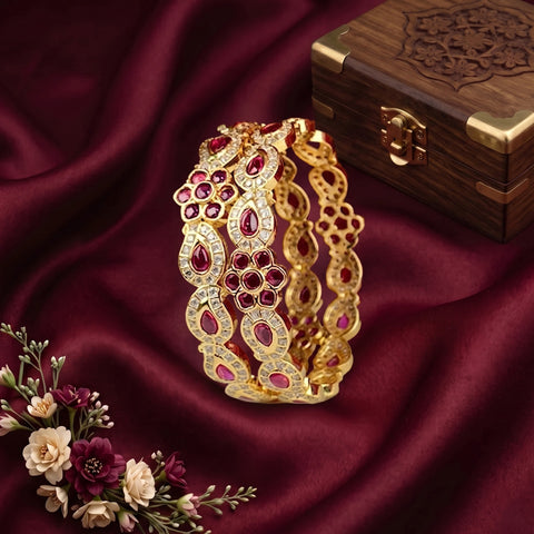 A gold-toned multi-strand bangle bracelet decorated with red gemstones and clear rhinestones, displayed on a draped burgundy satin fabric next to a wooden jewelry box and small flowers.