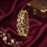 An ornate gold-toned traditional Indian bridal bangle bracelet decorated with green and clear stones, displayed on draped dark red satin fabric next to a wooden jewelry box and a small floral arrangement.