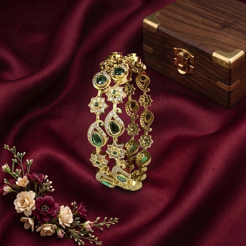 An ornate gold-toned traditional Indian bridal bangle bracelet decorated with green and clear stones, displayed on draped dark red satin fabric next to a wooden jewelry box and a small floral arrangement.