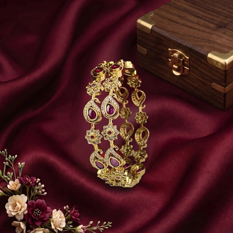 An ornate gold-toned bangle bracelet with teardrop-shaped purple gemstones and floral motifs, displayed on draped burgundy satin fabric beside a wooden box and small flowers.