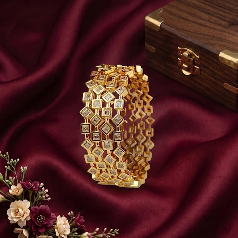 A gold-toned multi-strand bracelet with clear gemstones, displayed on draped burgundy satin fabric next to a wooden jewelry box and small flowers.