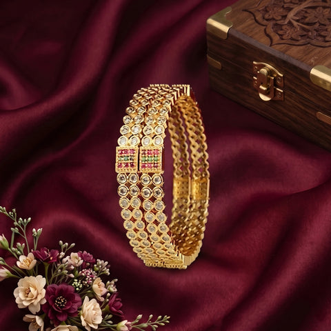 A gold-toned multi-strand bangle bracelet with square red and green stone accents, displayed on draped burgundy fabric next to a small floral arrangement and a carved wooden box.
