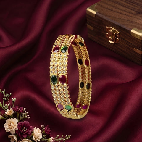 A gold-plated bangle bracelet with inlaid green and red stones and rows of clear crystals, displayed on draped burgundy satin fabric next to a wooden box and flowers.