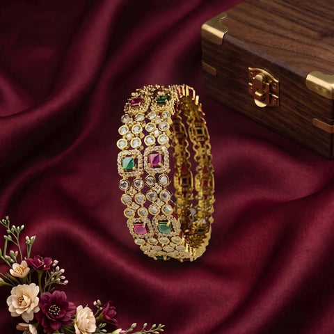 A gold-plated Indian-style bangle bracelet decorated with multiple rows of clear stones and green and pink gemstones, displayed on a draped dark red fabric next to a wooden jewelry box.