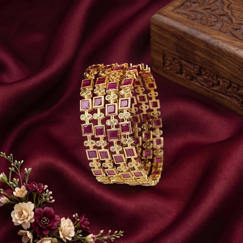 A gold-plated Indian style bangle bracelet with square purple gemstone accents, displayed on a draped dark red satin fabric next to a carved wooden box and flowers.