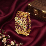 A gold-toned multi-strand bracelet set with square and floral motifs, embellished with pink and green stones, displayed on draped burgundy satin fabric next to a wooden jewelry box and decorative flowers.