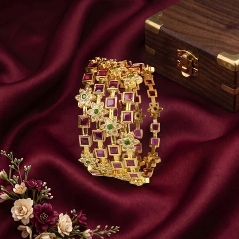 A gold-toned multi-strand bracelet set with square and floral motifs, embellished with pink and green stones, displayed on draped burgundy satin fabric next to a wooden jewelry box and decorative flowers.