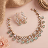 A gold-toned jewelry set featuring a necklace and matching drop earrings, decorated with clear stones and light green teardrop-shaped accents, arranged on a soft pink background with burlap and a flower prop.