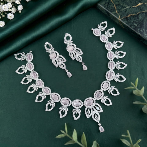 Silver-toned jewelry set with a necklace, earrings, and matching bracelet, featuring pale pink oval stones and intricate floral motifs, arranged on a dark green satin fabric with small white flowers and green leaves.