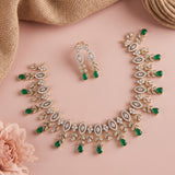 Gold-toned jewelry set with green teardrop gemstones and clear crystal-like stones, arranged on a soft pink background with burlap and a pink flower prop.