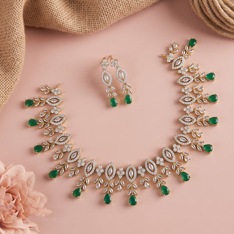 Gold-toned jewelry set with green teardrop gemstones and clear crystal-like stones, arranged on a soft pink background with burlap and a pink flower prop.