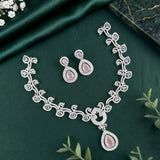 A silver-toned traditional Indian jewelry set featuring a necklace, earrings, and a bracelet, decorated with intricate floral patterns and pink teardrop-shaped stones, displayed on a dark green satin fabric background.