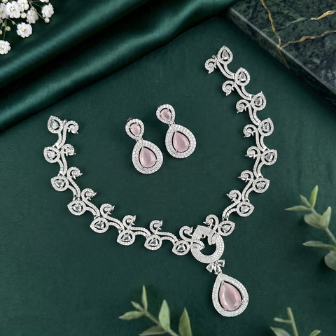 A silver-toned traditional Indian jewelry set featuring a necklace, earrings, and a bracelet, decorated with intricate floral patterns and pink teardrop-shaped stones, displayed on a dark green satin fabric background.
