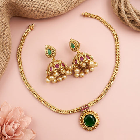 Gold-toned traditional jewelry set with a necklace and matching earrings, featuring green stones, pink stones, and pearl-like drops, arranged on a pink background with burlap and a flower prop.