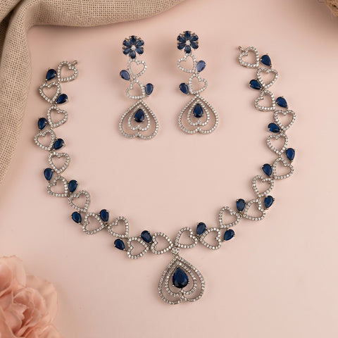 Silver-toned jewelry set with heart-shaped links, deep blue teardrop stones, matching drop earrings, and a teardrop pendant, arranged on a soft pink background with a beige fabric corner and a pale pink flower.