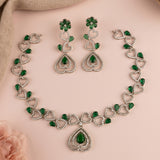 A silver-toned jewelry set featuring a heart-link necklace, matching drop earrings, and a teardrop pendant, decorated with green teardrop and flower-shaped stones, arranged on a light pink background.