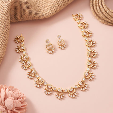 Gold-toned jewelry set with a necklace, matching earrings, and bracelet arranged on a soft pink background with burlap and a pink flower prop.